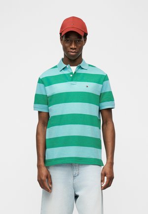Ung mand iført en grøn og lyseblå stribet polo-shirt, lyseblå jeans og en rød baseballkasket, stående mod en hvid baggrund.