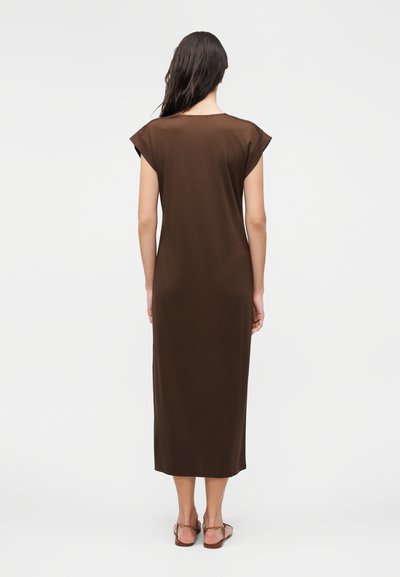 Γυναίκα με μακριά σκούρα μαλλιά που φοράει αμάνικο καφέ midi φόρεμα και ίσια σανδάλια, στέκεται γυρισμένη με την πλάτη σε απλό φόντο.
