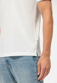 Witte polo shirt van een gestructureerde stof, met een rechte zoom, zijsplitten en blauwe accentstiksels langs de kraag en mouwen.