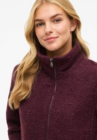 Blonde sieviete ar gariem matiem, kas smaida, tērpusies tumši violetā augsta apkakle, ar rāvējslēdzēju un faktūru fliša jakā.