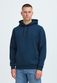 Marineblå hettegenser laget av mykt materiale, med en frontlomme, justerbare snorer og en liten svart logo på brystet.