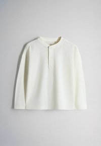 Camiseta henley blanca de manga larga con dos botones, cuello redondo y corte relajado sobre un fondo gris claro liso.