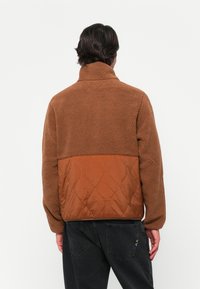 Brown fleece jacket with a high collar, textured body, and contrasting quilted panel at the lower back. Features a relaxed fit and long sleeves.