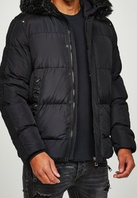 Muž v černé prošívané bundě s kapucí lemovanou kožešinou, černém tričku a potrhaných černých džínách, stojící s pažemi volně podél těla.