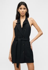 Γυναίκα που φοράει αμάνικο μαύρο φόρεμα-σακάκι με ζώνη, μπροστινές τσέπες, λεπτομέρειες με κουμπιά και βαθύ ντεκολτέ σε σχήμα V, στέκεται με το ένα χέρι στη μέση.