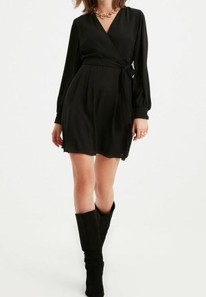 Vrouw draagt een zwarte wikkeljurk met lange mouwen gestrikt in de taille, zwarte kniehoge laarzen en een stevige gouden ketting.