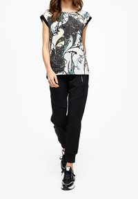 Top grafico con maniche corte in bianco, nero e design turchese; abbinato a pantaloni jogger neri; indossato da un modello con sneakers nere.
