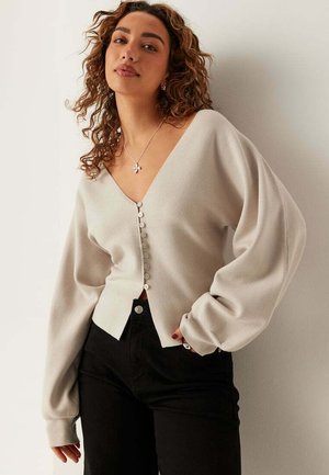 Kvinde med krøllet hår iført en beige cardigan med knapper og puffede ærmer samt sorte bukser, stående op ad en hvid væg.