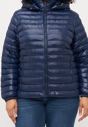 Frau trägt eine marineblaue gesteppt gefütterte Jacke mit Kapuze, Reißverschluss vorne und Seitentaschen, kombiniert mit blauen Jeans.