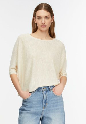 Frau mit langen glatten Haaren, die einen cremefarbenen, locker gestrickten Pullover und hellblaue, hoch taillierte Jeans trägt, die Hände in den Taschen.