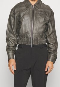 Man med en kort grå läderjacka med dragkedja fram, långa ärmar och högmidjade svarta byxor mot en neutral bakgrund.