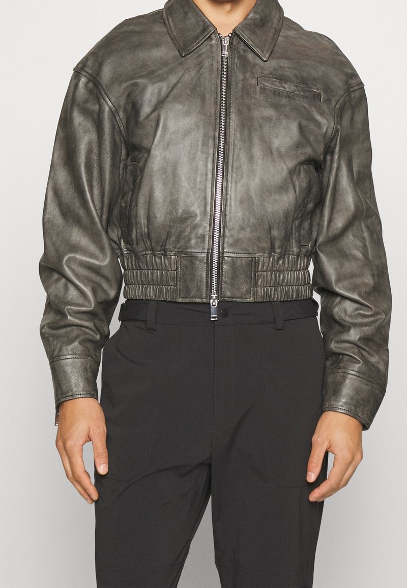 Man med en kort grå läderjacka med dragkedja fram, långa ärmar och högmidjade svarta byxor mot en neutral bakgrund.