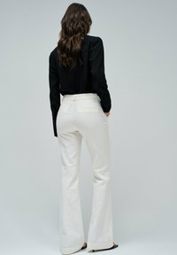 Pantalones acampanados blancos con una textura suave y bolsillos traseros simples, combinados con una blusa negra de manga larga. Fondo neutro.
