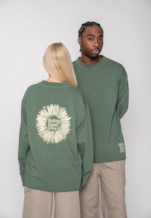 Dvaja ľudia oblečení v zelených tričkách s dlhým rukávom a béžových nohaviciach, jeden stojí čelom a druhý ukazuje chrbát trička s bielym kruhovým textovým dizajnom.