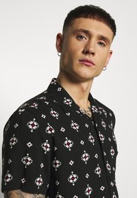 Korte mouwen zwarte shirt met een geometrisch patroon in het wit en rood, met een klassieke kraag en knoopsluiting aan de voorkant. Gladde textuur.