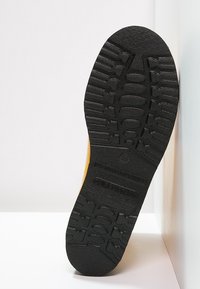 Suela de goma negra con un patrón texturizado. Material superior amarillo visible a lo largo del borde, mostrando el número de talla 39.