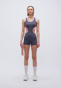 Νεαρή γυναίκα με αθλητική ολόσωμη φόρμα σε σκούρο γκρι και ροζ με ανοίγματα, κρατώντας ροζ λουρί, φορώντας λευκές κάλτσες και αθλητικά παπούτσια, στέκεται σε λευκό φόντο.