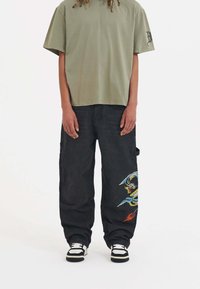 Olivgrön kortärmad t-shirt i kombination med svarta cargobyxor som har färgglad drakbroderi och en avslappnad passform. Svartvita sneakers.