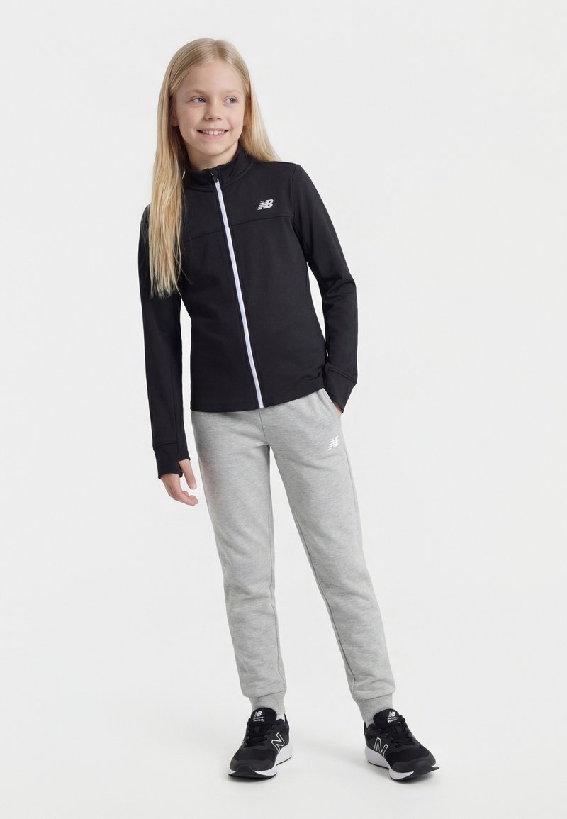 Κορίτσι με μακριά ξανθά μαλλιά, που φοράει μαύρο μπουφάν με φερμουάρ, γκρι φόρμες και μαύρα αθλητικά παπούτσια, στέκεται μπροστά σε λευκό φόντο.