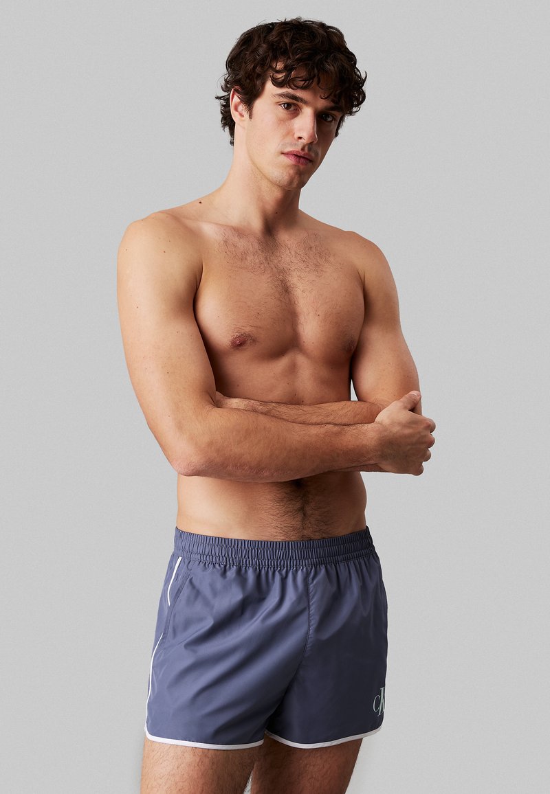 Pantaloni da bagno grigi scuri con vita elasticizzata, rifiniture bianche e dettaglio del logo sul davanti. Texture liscia e silhouette aderente.