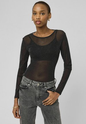 Femme portant un haut noir pailleté transparent à manches longues sur un bralette noir et un jean noir délavé taille haute, la main dans la poche.