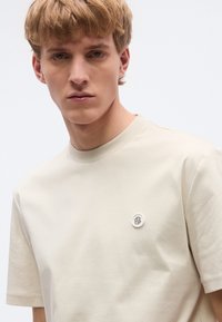 Μπεζ κοντομάνικο T-shirt κατασκευασμένο από βαμβάκι, με στρογγυλή λαιμόκοψη και μικρό στρογγυλό λογότυπο στο στήθος. Ομαλή υφή.