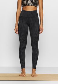 Czarne legginsy z teksturowanym, paskowym wzorem. Wysoki stan, dopasowany krój. Stopą bez butów, stojąca na jasnej podłodze.