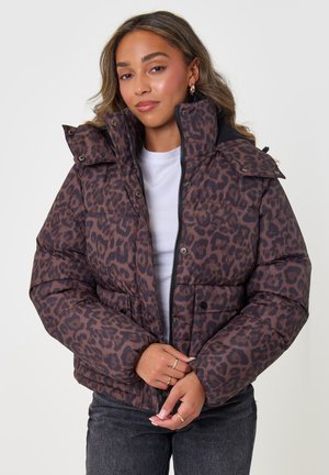 Mujer con una chaqueta acolchada marrón y negra con estampado de leopardo sobre una camisa blanca y vaqueros oscuros contra un fondo liso y claro.