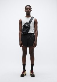 Μαύρο σορτς για τρέξιμο, γκρι αμάνικο τοπ, μαύρη τσάντα χιαστί με λογότυπο και αθλητικά παπούτσια με χρυσές και κόκκινες λεπτομέρειες.