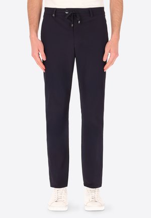 Uomo che indossa pantaloni neri slim fit con tasche laterali e sneakers bianche, in piedi contro uno sfondo semplice.