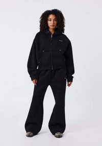 Sudadera negra de mezcla de algodón con cremallera frontal y dos bolsillos delanteros. Combinada con pantalones acampanados a juego. La modelo está de pie frente a un fondo neutro.