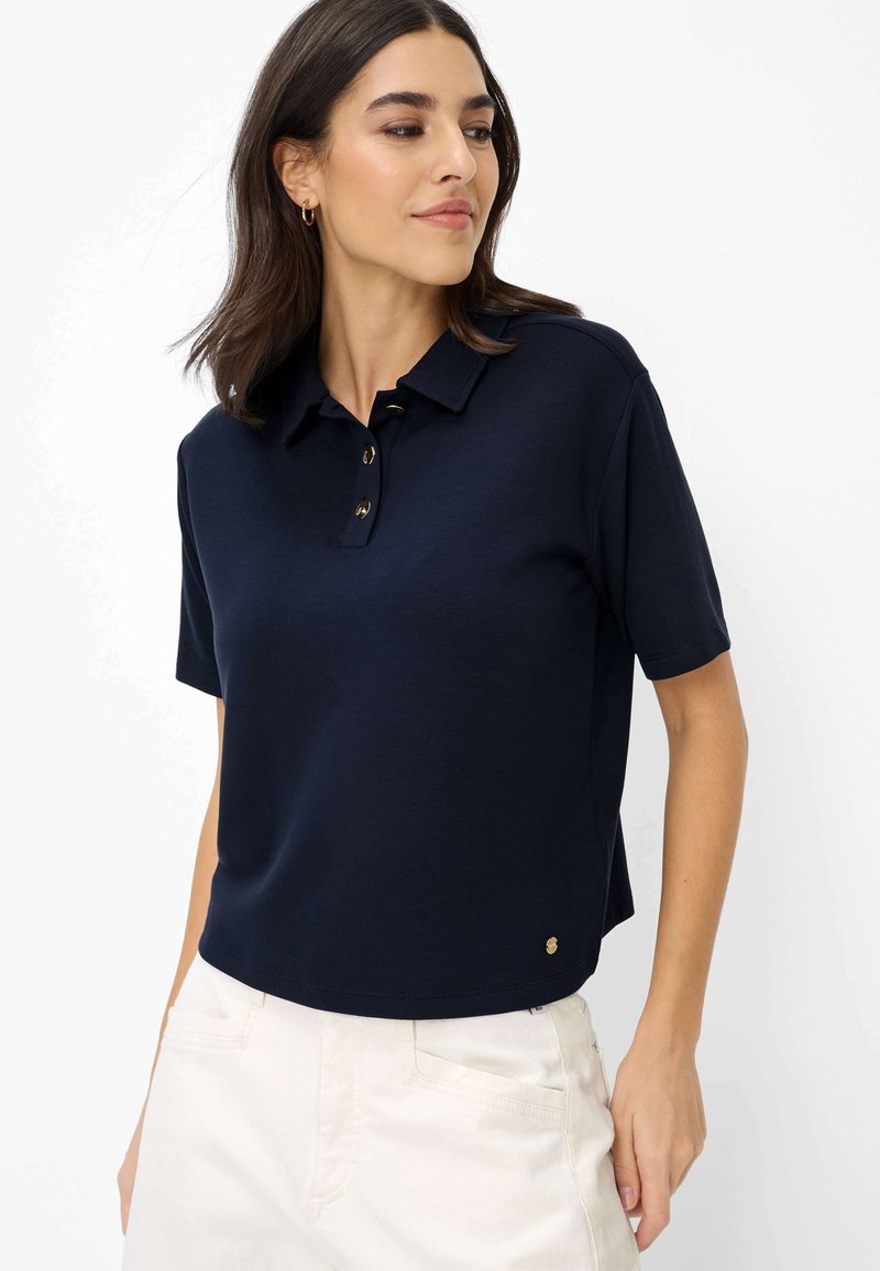 Femme portant un polo bleu marine à manches courtes avec des boutons, accompagné d'un pantalon blanc, regardant légèrement sur le côté sur un fond blanc.