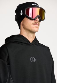 Haut à capuche de snowboard noir avec une finition texturée, arborant un emblème de logo, porté avec un bonnet en tricot noir et des lunettes de ski miroitées avec un teinte réfléchissante.