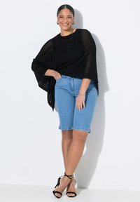 Poncho negro de punto abierto sobre shorts de denim azul claro, con un ajuste relajado y costuras visibles. Sandalias de tacón con tiras negras completan el look.