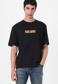 Svart t-skjorte med kort arm som har "HUGO MANIA" i fet gul tekst foran, sammen med lyseblå jeans. Glatt stoffstruktur.