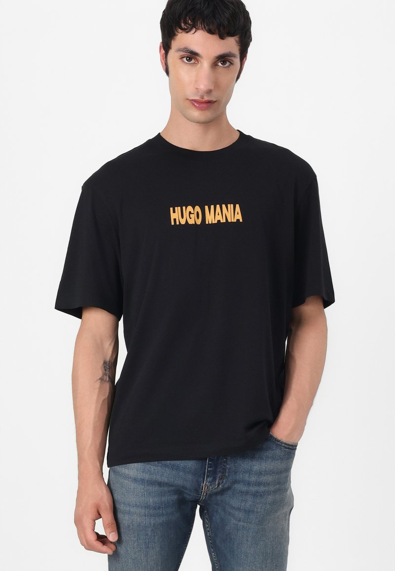 Czarny t-shirt z krótkim rękawem z napisem "HUGO MANIA" w wyrazistym żółtym kolorze z przodu, w połączeniu z jasnoniebieskimi dżinsami. Gładka faktura materiału.