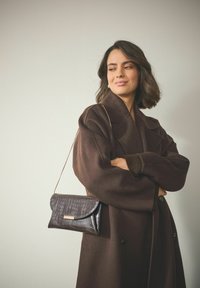 Schwarze Clutch mit Krokodilprägung, Klappdesign und goldfarbenen Hardware-Akzenten. Kombiniert mit einem braunen Wollmantel.