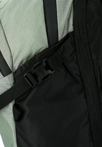 Primer plano de una cremallera negra y una correa ajustable con hebilla de plástico en un panel de tela de mochila gris y negra.