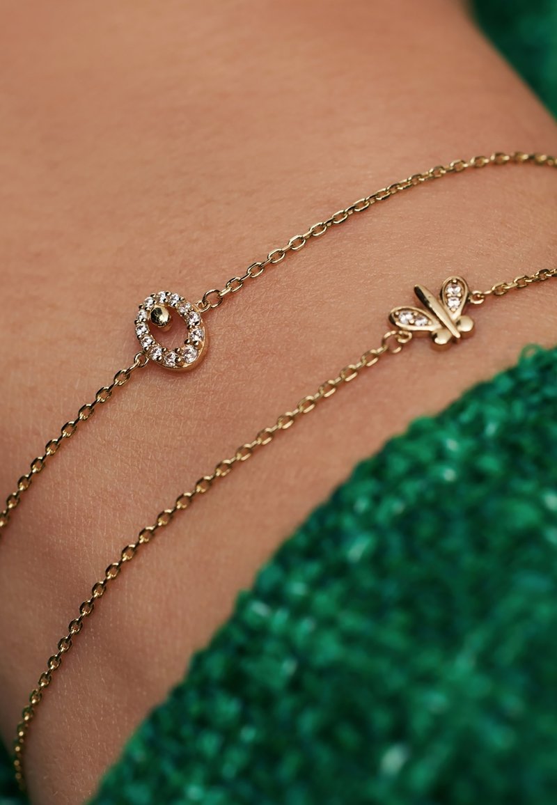 Kultaketju rannekoru, jossa on kaksi riipusta: yksi ovaali, jossa on kirkkaat kivet, ja toinen perhosen muotoinen, molemmat kiillotetulla pinnalla.