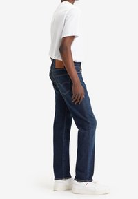 Jeans de denim azul oscuro con un corte recto, que presentan una etiqueta de cuero marrón en la cintura y detalles de costura sutiles. Combinados con una camiseta blanca.