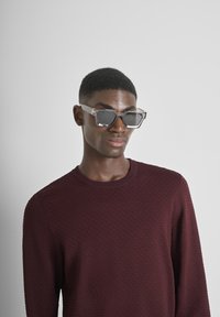 Occhiali da sole trasparenti a forma esagonale con lenti scure. Modello che indossa un maglione texturizzato bordeaux. Sfondo morbido in grigio chiaro.