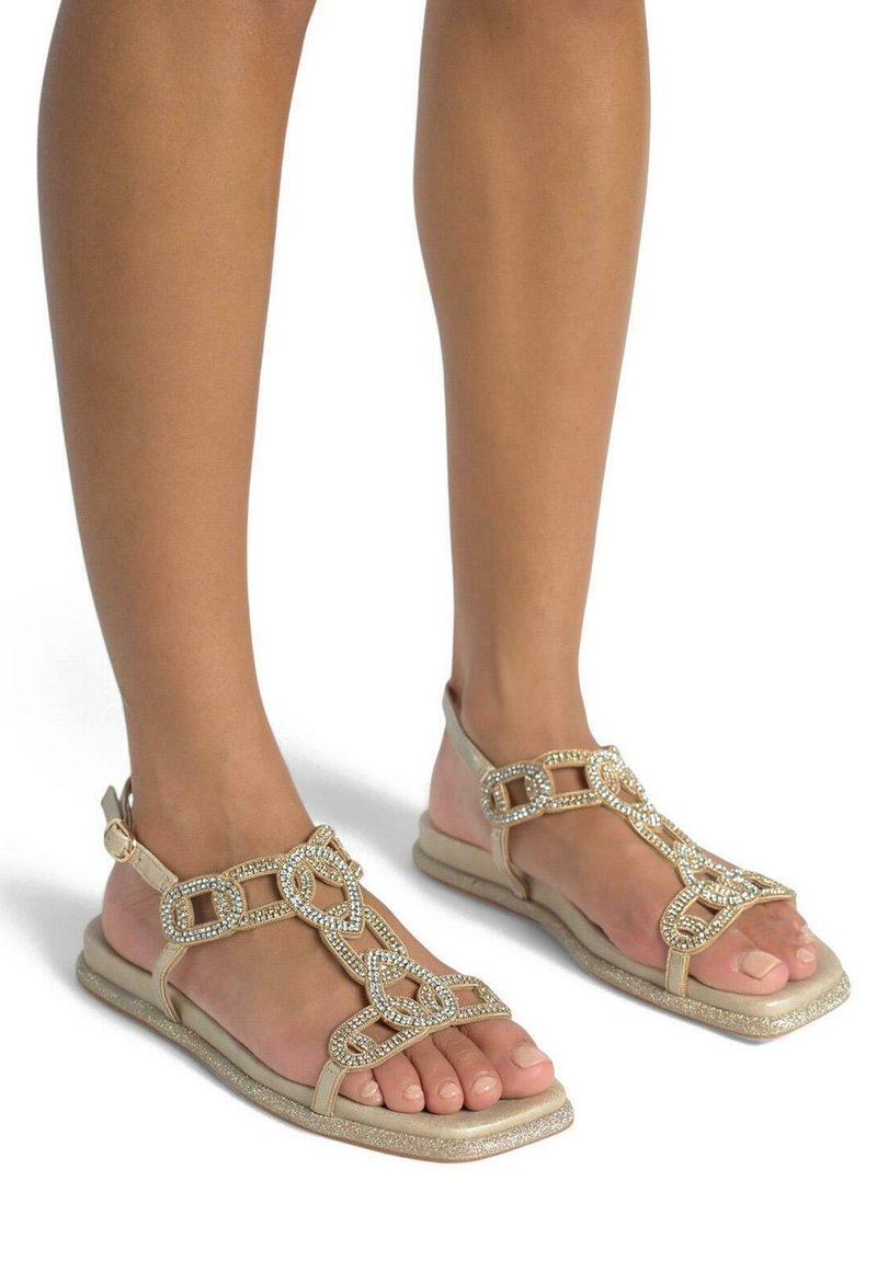 Sandales plates dorées présentant un design T-strap orné de bijoux avec des maillons en chaîne et une semelle plate texturée. Style ouvert à l'avant mettant en valeur la pédicure.