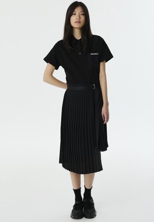 Zwarte polo met opgerolde mouwen, gecombineerd met een zwarte plissérok en een bijpassende riem. Draag het met zwarte sneakers en scheenSokken.