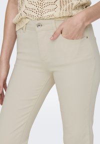 Primo piano di pantaloni a vita alta crema con bottone e zip, una mano in tasca, indossati con un top in crochet beige.