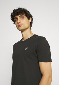 Black cotton t-shirt featuring a round neck and short sleeves, with a small white bird logo embroidered on the left chest.