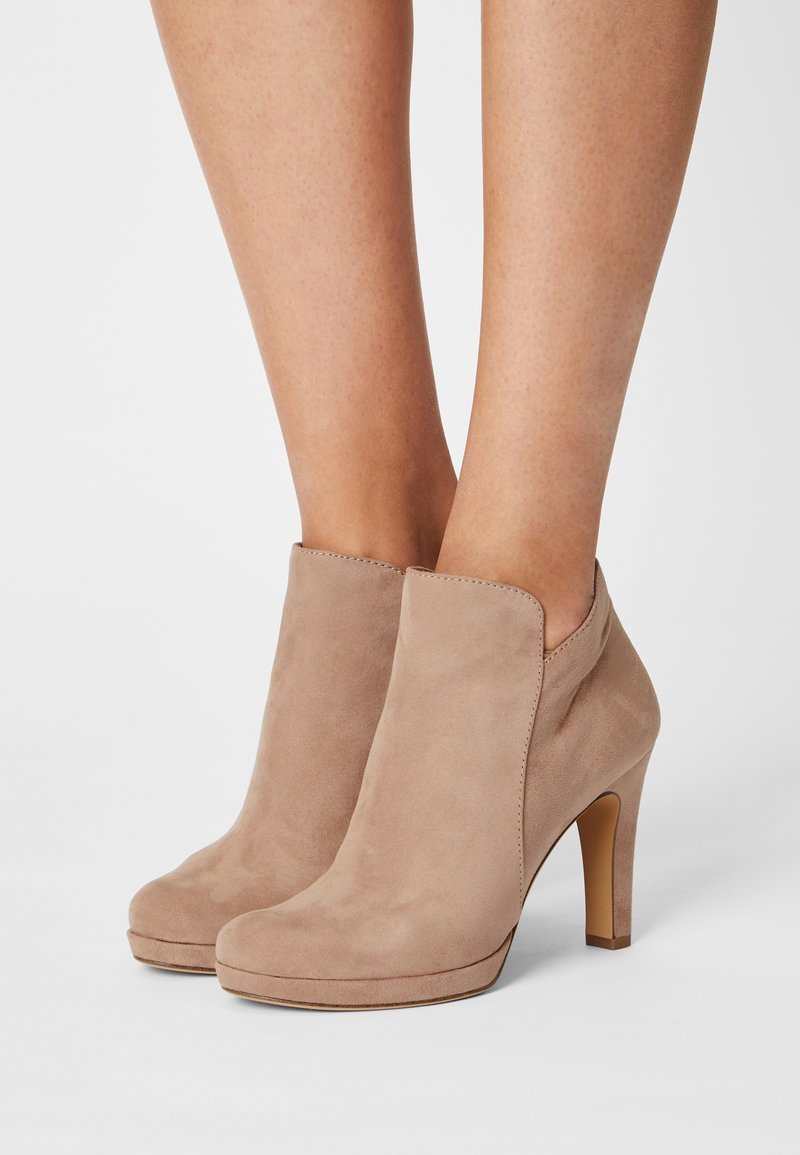 Jambes portant des bottines beiges en daim à talons hauts avec bouts arrondis sur fond blanc.