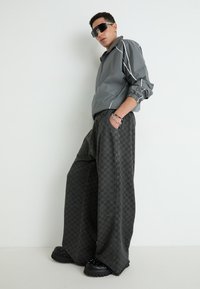 Gray windbreaker with white accents, loose black checkered pants, and chunky black shoes with studs. Model is wearing oversized sunglasses.