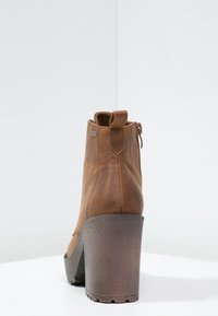 Brązowy skórzany botek do kostki z wysokim, teksturowanym obcasem i suwakiem z tyłu. Gładkie wykończenie z pętelką ułatwiającą zakładanie.