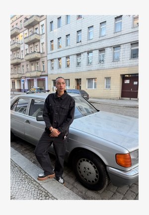 Noor naine mustas riietuses toetub hõbedasele vintage-autole, mis on pargitud munakiviteel linnalähedases elamupiirkonnas.