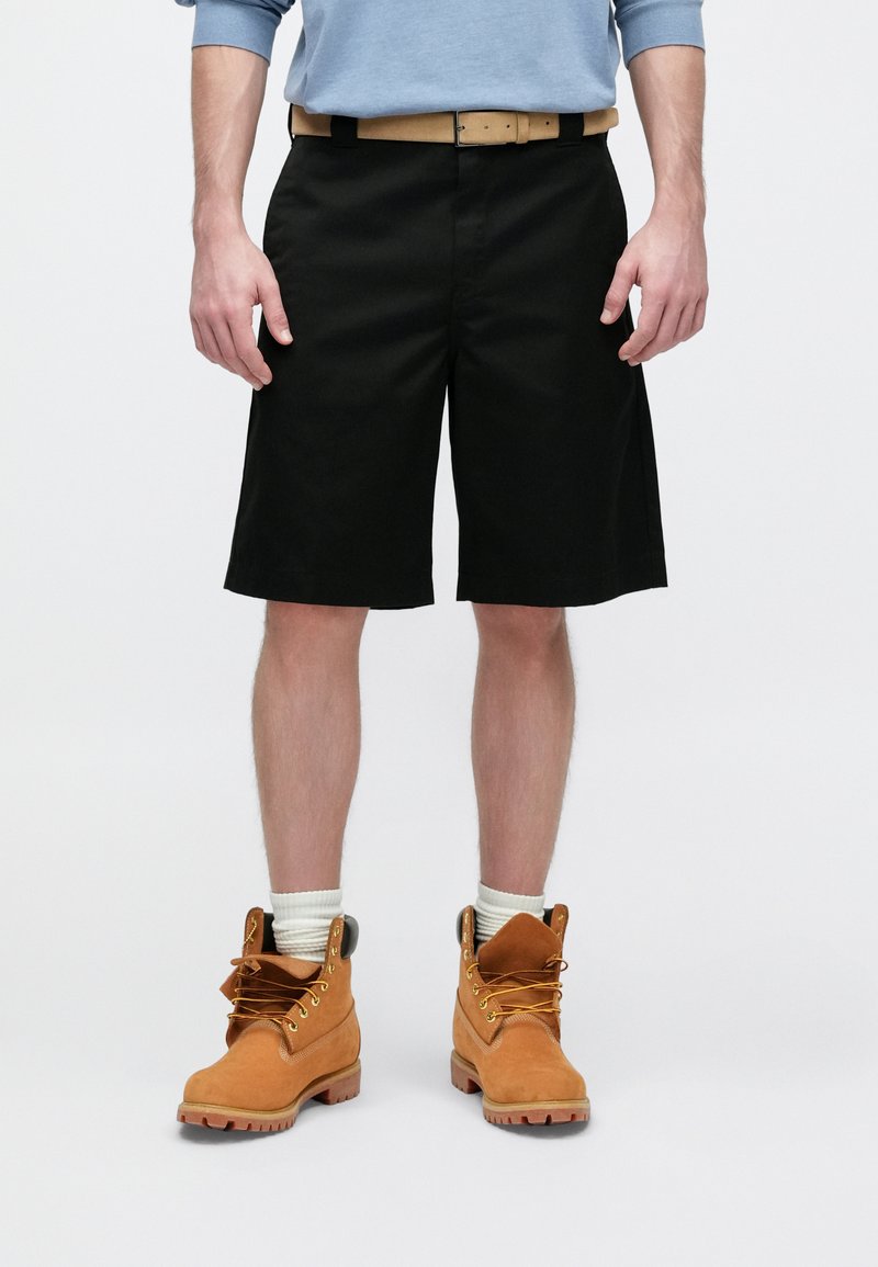 Pantaloncini neri a vestibilità ampia, una cintura in pelle marrone chiaro e stivali in camoscio arancione. Gli stivali hanno lacci e imbottitura intorno alle caviglie.